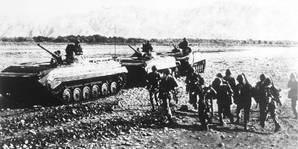 Black and white wartime scene with infantry moving beside tracked armored vehicles on a rocky riverbank as crew members sit on top with mounted guns.