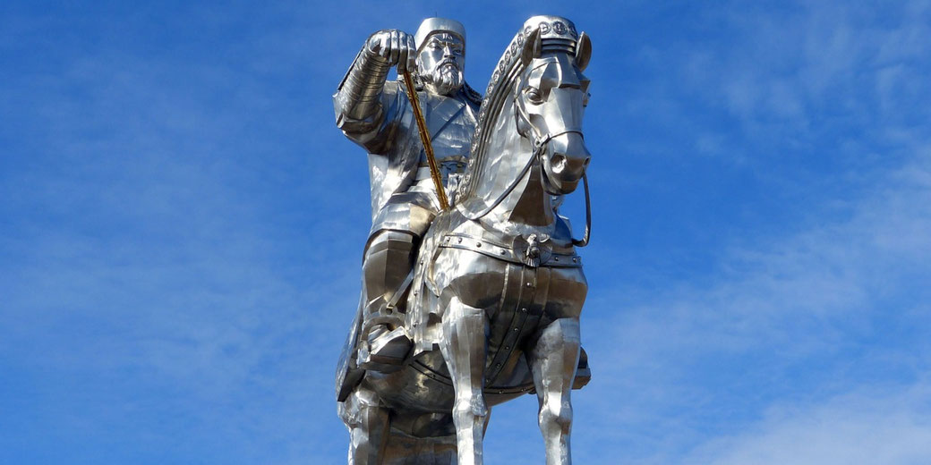 A massive silver statue of a warrior on horseback, made of reflective metal, set against a bright blue sky. The figure holds a golden whip and wears traditional armor and a hat.