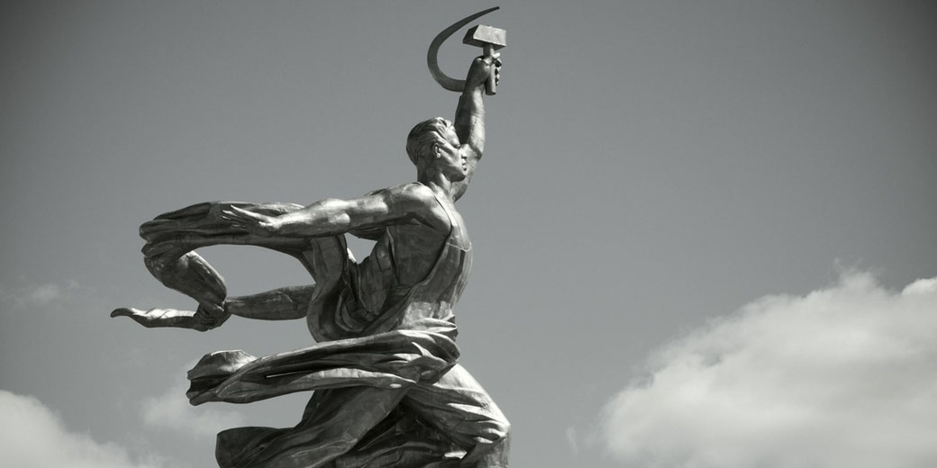 Dramatic metal statue of a man striding forward with raised arm holding a hammer and sickle, set against a partly cloudy sky.