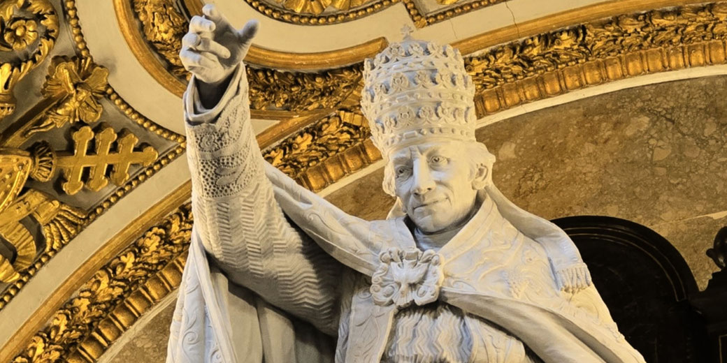 A marble statue of a pope in detailed papal robes and tiara, raising one hand, set beneath an arch with intricate gold embellishments.