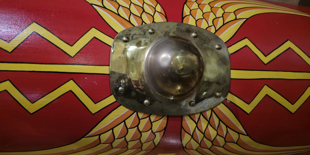 Close-up of a Roman shield's brass boss, surrounded by yellow wing and lightning bolt designs on a red background.