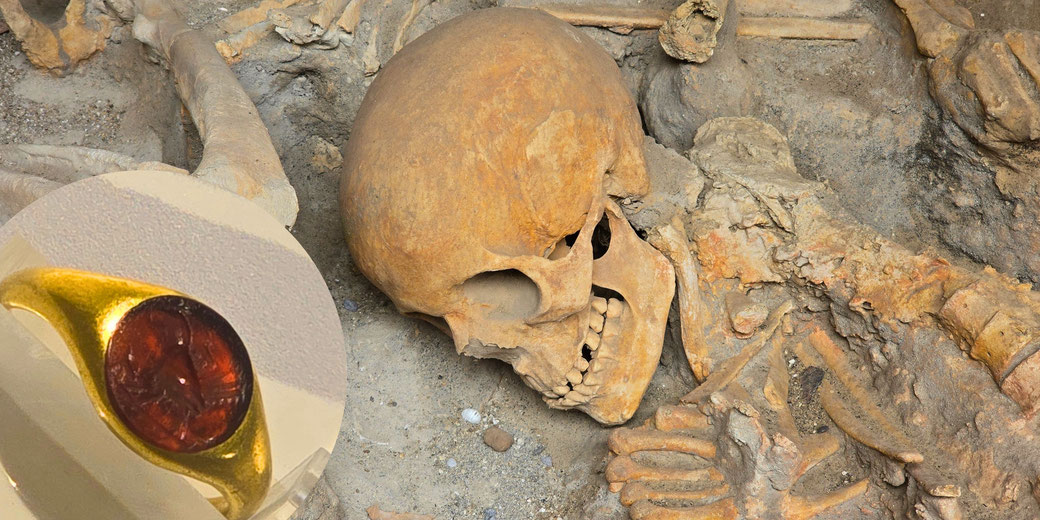 A human skeleton covered in volcanic ash lies amid scattered bones, its skull tilted to reveal teeth alongside a gold ring set with a deep red gemstone.