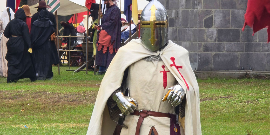 A person dressed as a medieval knight in a white cloak and steel helmet stands at a historical reenactment event with others in period clothing.