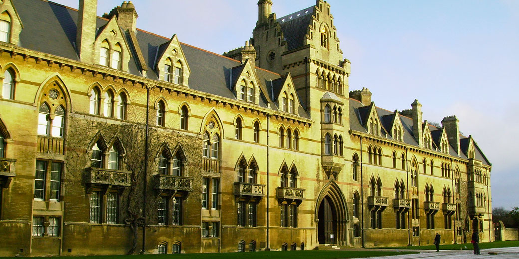 A grand Gothic-style building with arched windows, intricate stonework, and a towering entrance. The golden-hued facade is illuminated by sunlight, with a lawn and people walking nearby.