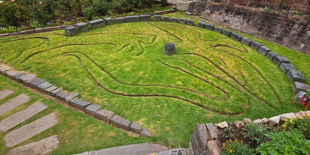 An ancient Incan earth carving depicting a stylized animal figure, possibly a puma or condor, outlined in grass and stone within an archaeological site surrounded by gardens.