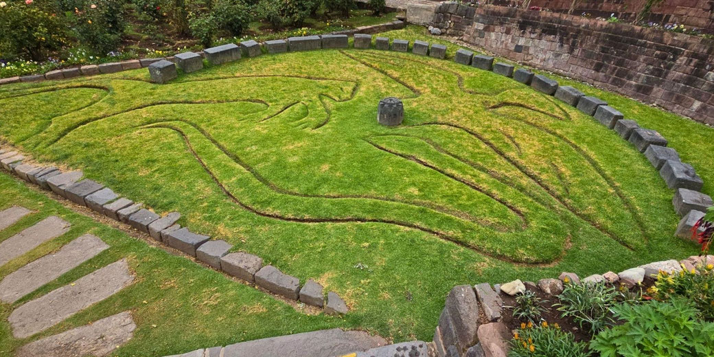 An ancient Incan earth carving depicting a stylized animal figure, possibly a puma or condor, outlined in grass and stone within an archaeological site surrounded by gardens.