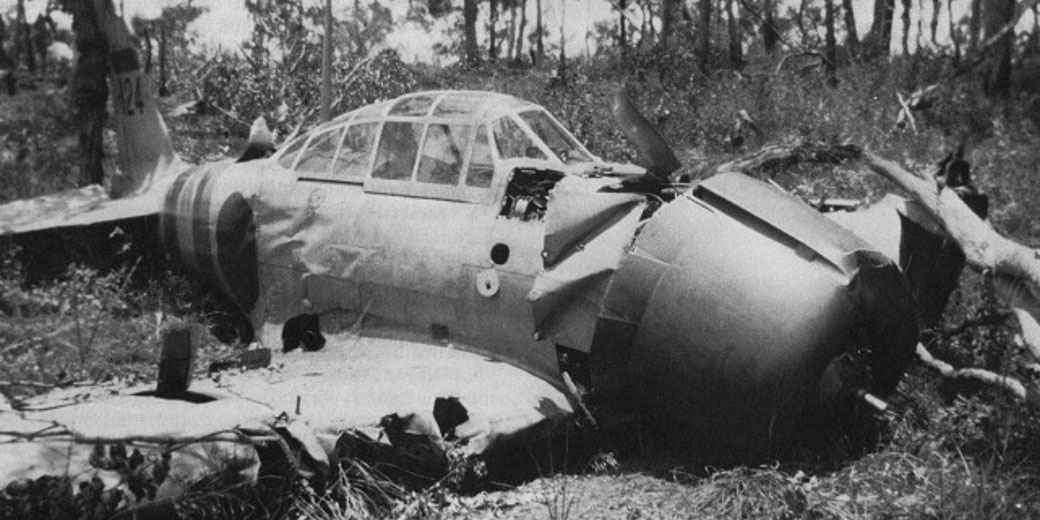A crashed WWII-era fighter plane lies wrecked in a forest clearing, with visible damage to the nose, cockpit, and wings.