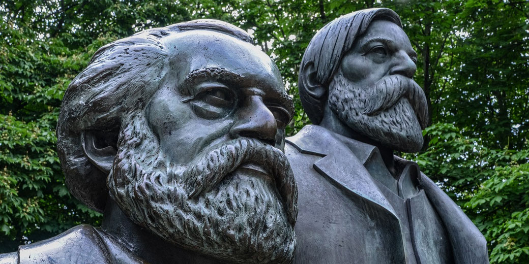 Close-up of a bronze statue depicting two bearded men with serious expressions, set against a backdrop of leafy green trees.