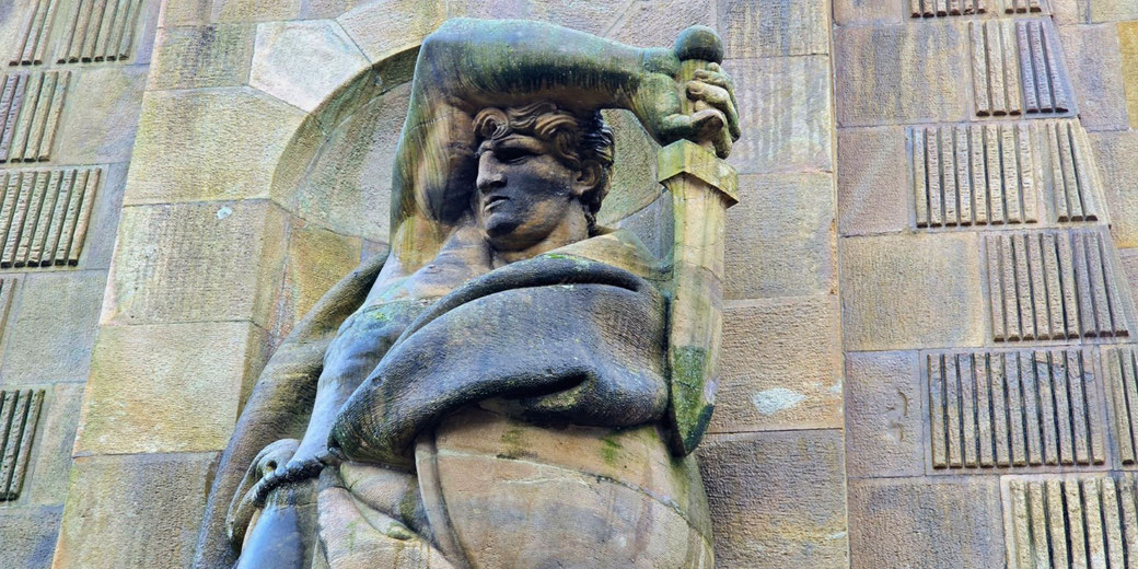 Stone statue of a muscular figure with a stern expression holding a sword overhead, set against a patterned stone wall.