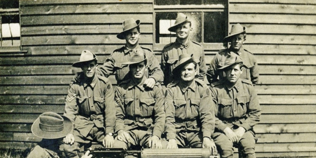 Group of World War I-era soldiers in uniform and slouch hats posing in front of a wooden building with a machine gun.