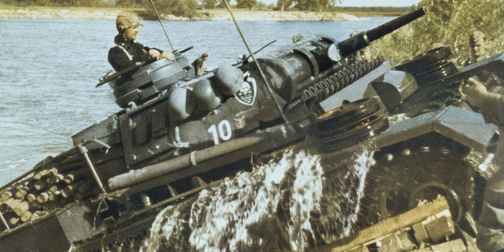 A military tank with soldiers crosses a river, water splashing off its sides as it climbs onto land, showing active wartime movement.