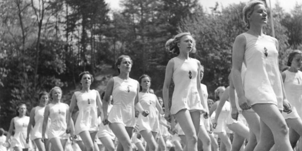 Black and white image of young women marching in matching athletic uniforms during an outdoor event with trees behind them.