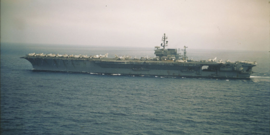 A large aircraft carrier sails through open ocean waters with numerous jets and helicopters visible on its deck.