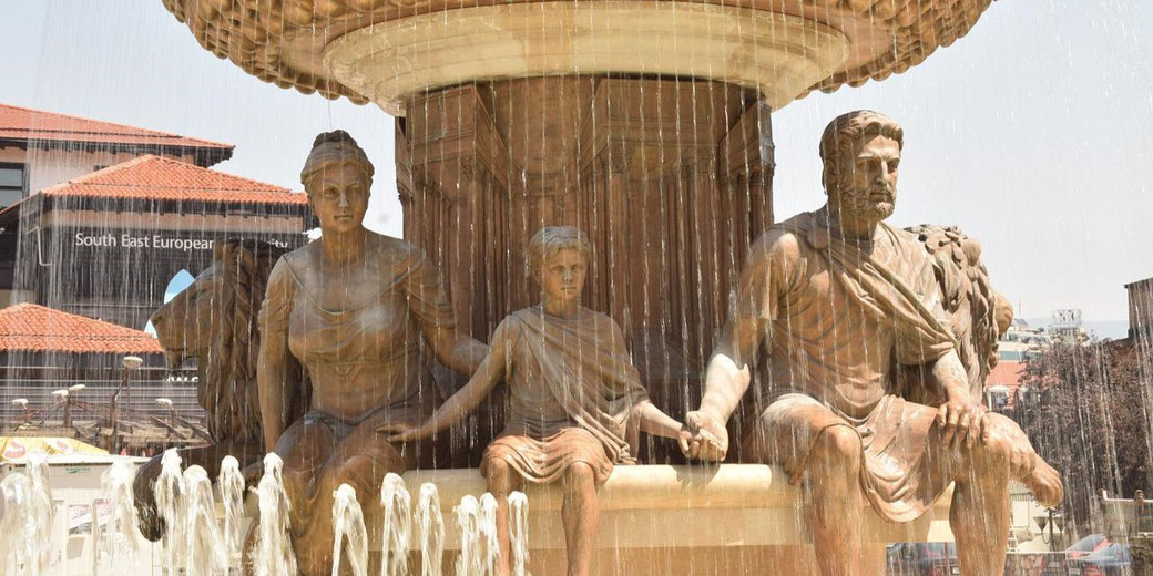 Bronze statues of a seated family, mother, father, and child, hold hands beneath cascading water in a large public fountain.