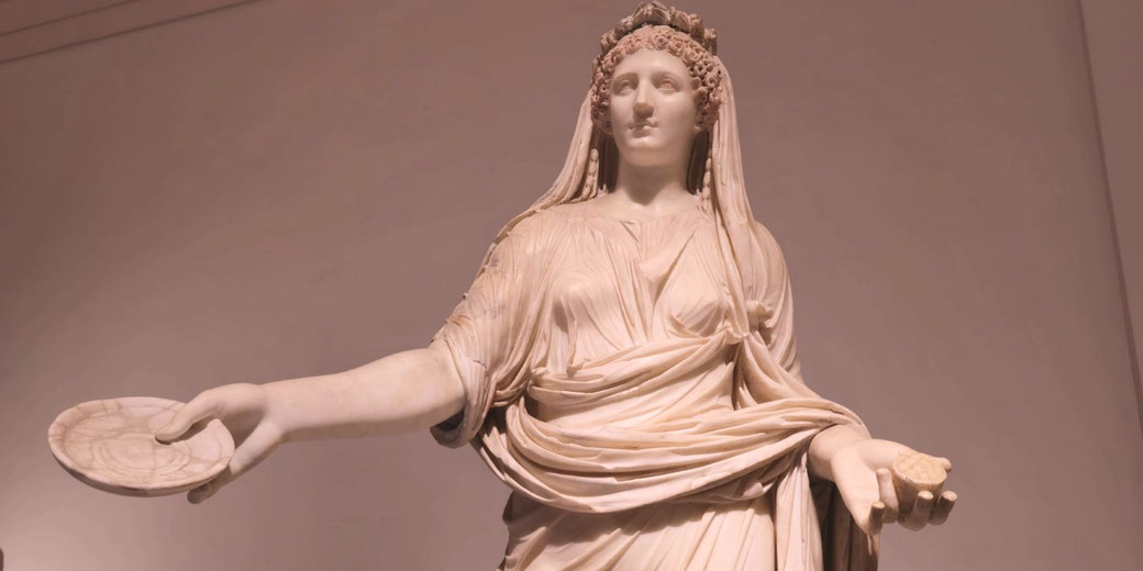 Marble statue of a veiled woman in flowing robes, holding a shallow dish in one hand and an offering in the other, likely representing a Roman goddess or priestess.