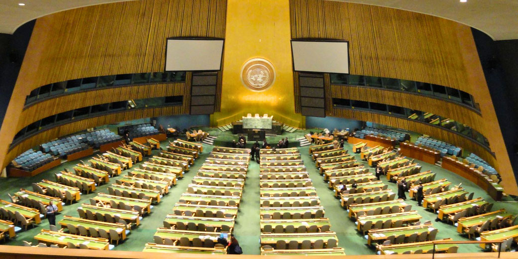 The United Nations General Assembly hall, featuring rows of seats, a central podium, and the UN emblem on a golden backdrop, set within a curved, wood-paneled chamber.