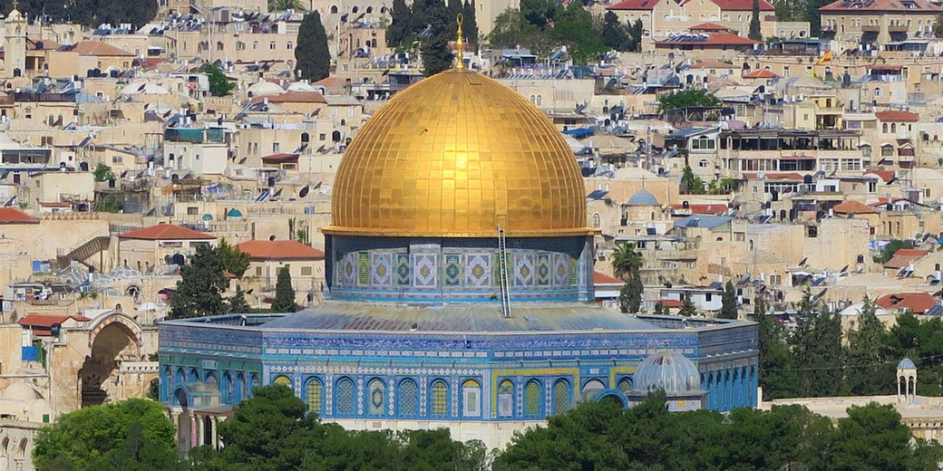 A golden dome sits atop a blue-tiled structure, surrounded by dense stone buildings and trees in an ancient city.