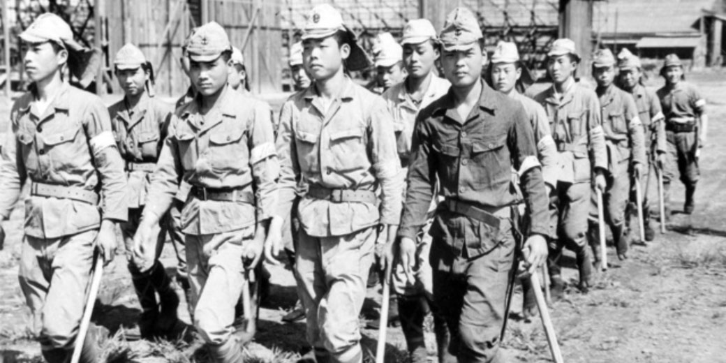 Group of Japanese soldiers in uniform march in formation holding rifles, likely during World War II, with wooden structures visible in the background.