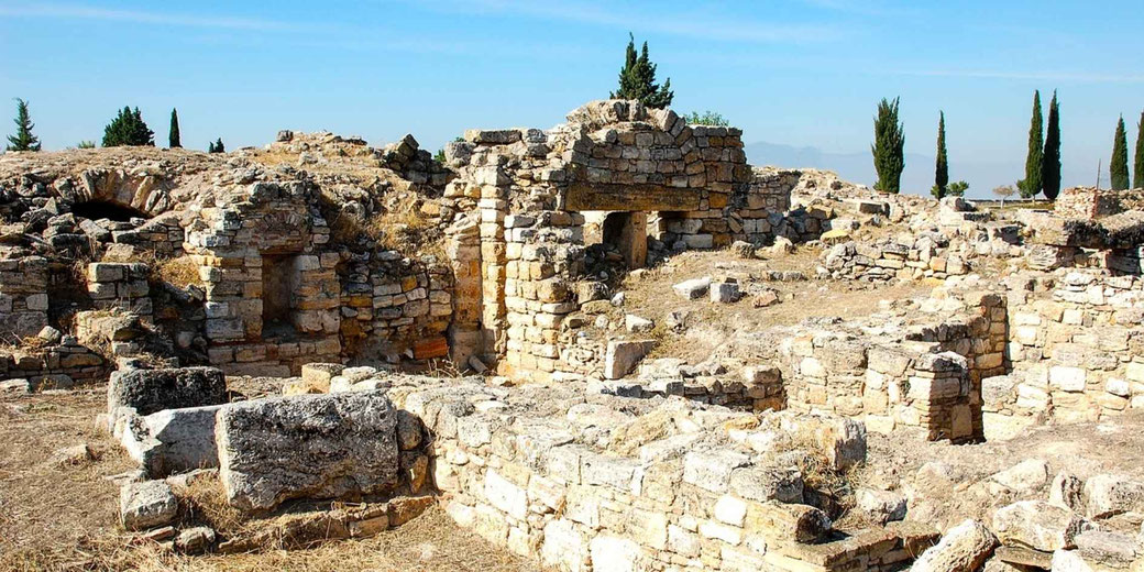 Ancient stone ruins with partially standing walls and archways under clear sky, surrounded by dry ground and sparse trees in the distance.