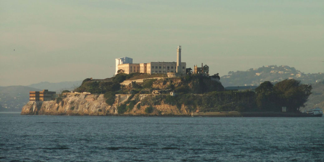An island with a large historic prison building and lighthouse rises from rocky cliffs, surrounded by water with distant hills in the background.