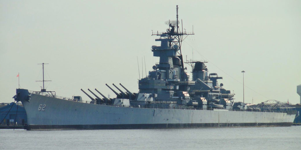 A large U.S. battleship with the number 62 is docked at port, showcasing multiple heavy gun turrets.