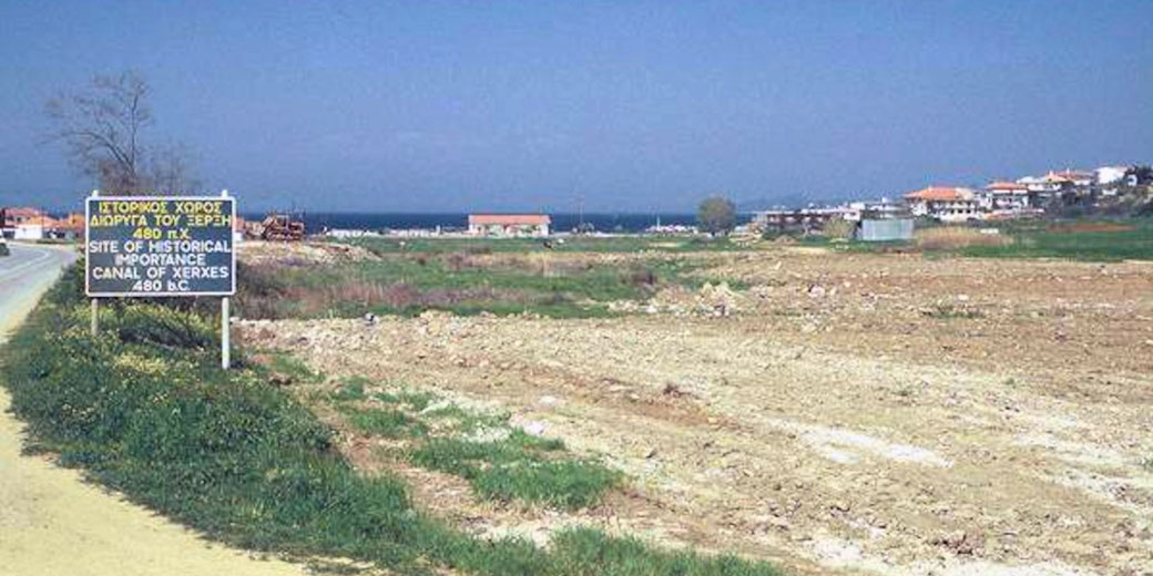 A roadside sign marks the Canal of Xerxes historical site near an open field with distant houses and the sea.