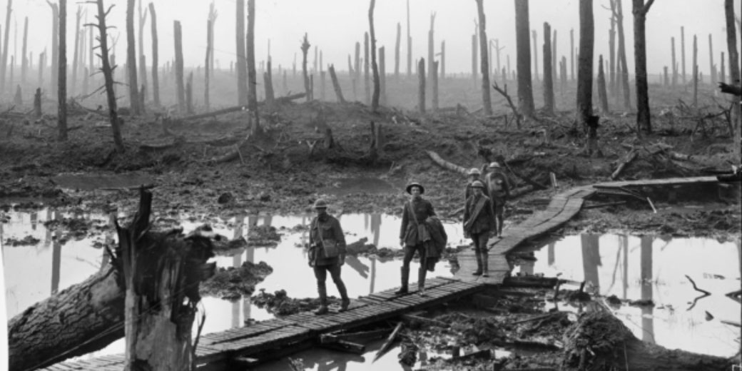 Soldiers walk across a narrow wooden path over waterlogged, muddy terrain surrounded by shattered trees in a World War I battlefield.