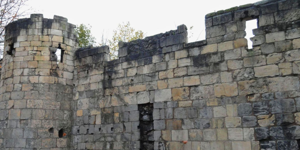 A close-up of a weathered stone castle wall with a round tower. The stones are uneven, with some missing or damaged, and there are small gaps or windows along the structure.