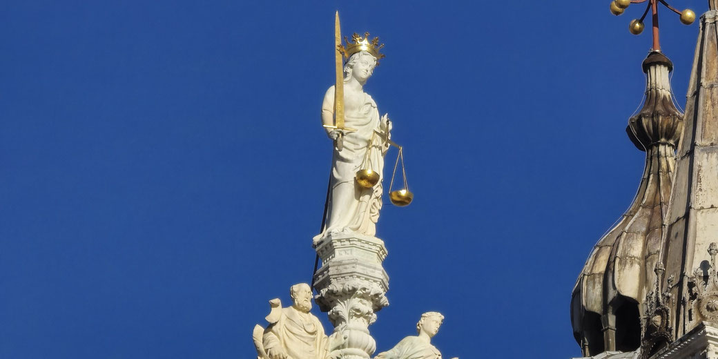 A statue of Lady Justice atop a historic building, holding golden scales and a sword. She wears a crown, symbolizing fairness and authority, against a clear blue sky.