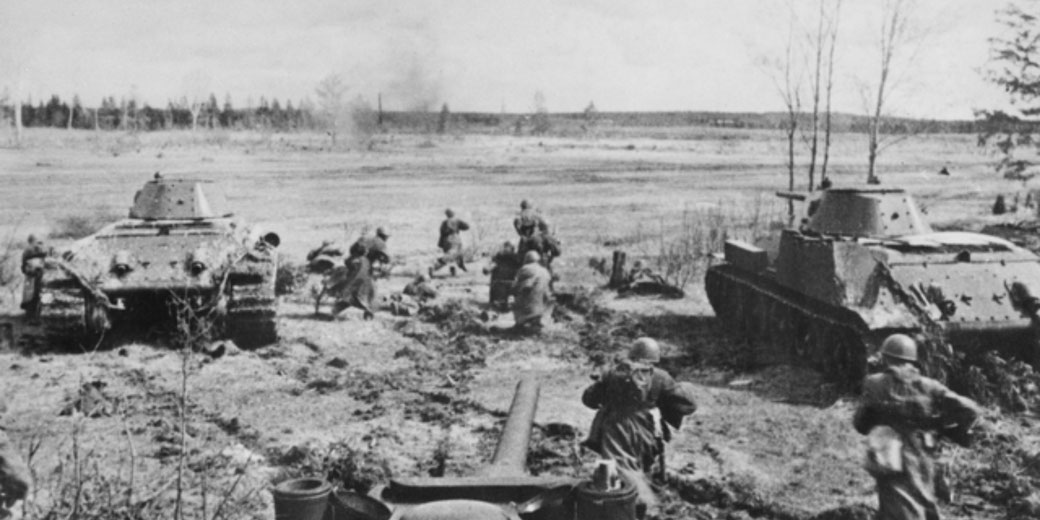 Soldiers advance between damaged tanks across a battlefield, with artillery positioned in the foreground and open terrain stretching behind them.
