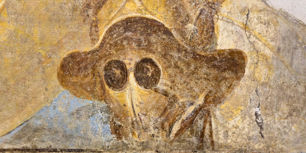 Ancient fresco showing a figure with a wide-brimmed hat and large round eyes, resembling a plague doctor mask, with faded colors and worn texture.