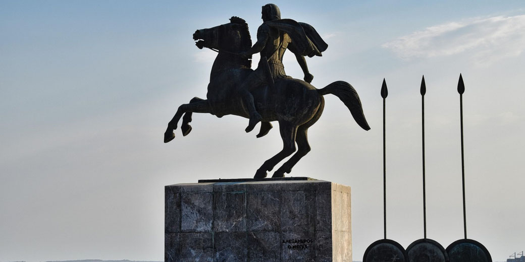 A bronze statue of Alexander the Great on horseback stands beside tall spears and round shields, facing an open sky.