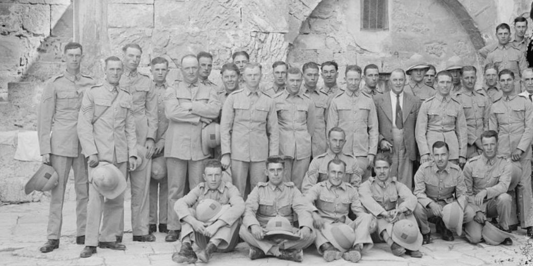 A group of uniformed soldiers poses for a photo in front of a stone wall, some seated and others standing in neat rows.