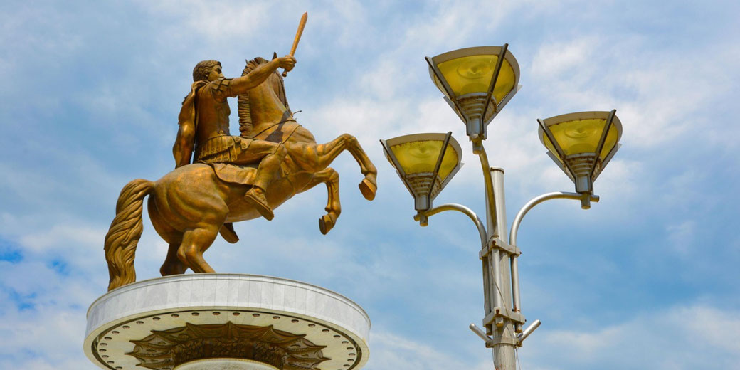 A bronze warrior on horseback raises a sword beside a tall streetlamp under a bright, partly cloudy sky.