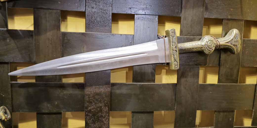 A Roman-style short sword with a wide, double-edged blade, central ridge, and a decorated hilt featuring engraved patterns and a rounded pommel.