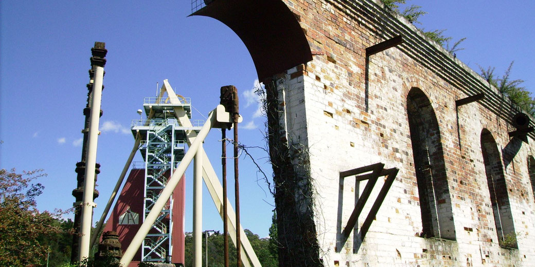 An old brick industrial structure with arched windows is partially covered in vines. A modern metal mining headframe and rusted machinery stand nearby under a clear blue sky.