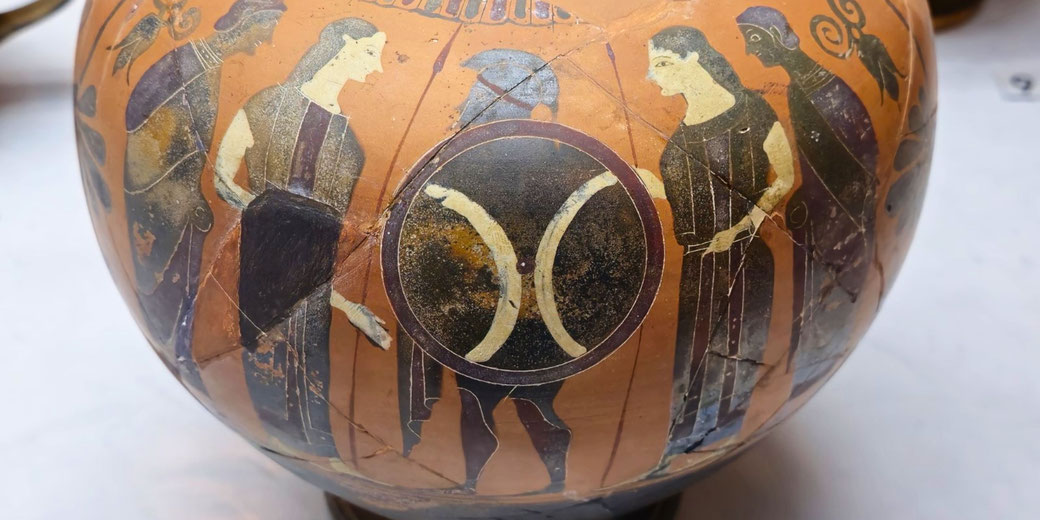 Ancient Greek ceramic vase featuring black-figure artwork of warriors and women, with a central shield motif and visible aging cracks.