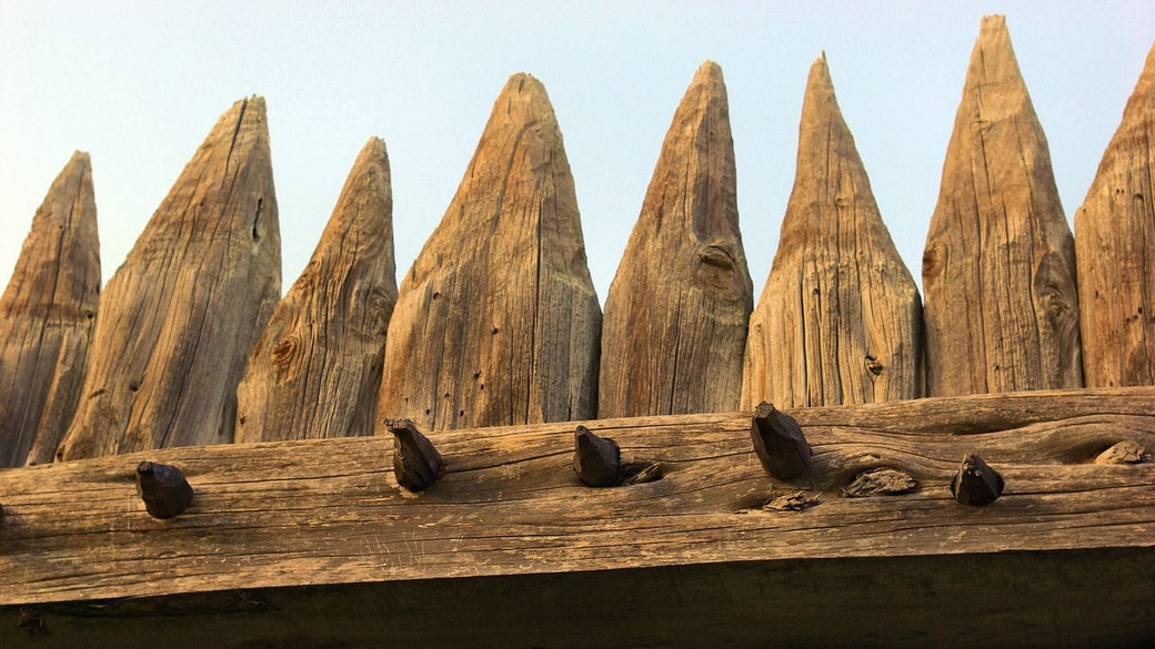 A close-up of a wooden palisade with sharp, pointed stakes and visible fasteners against a clear sky.