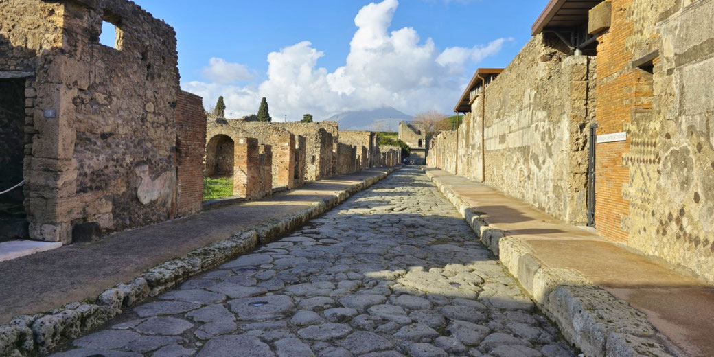 Stone-paved ancient Roman street in Pompeii lined with ruins, leading toward Mount Vesuvius under a partly cloudy sky.