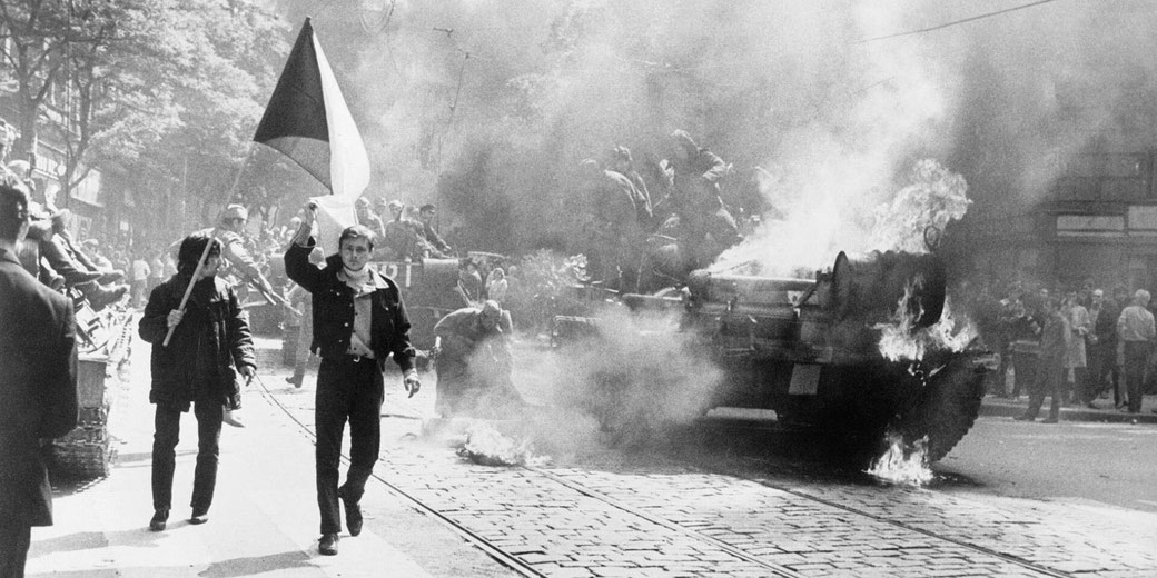 Black and white photo of a protest scene with a man holding a flag walking toward a burning tank as smoke rises. Crowds watch from the street during a tense urban uprising.