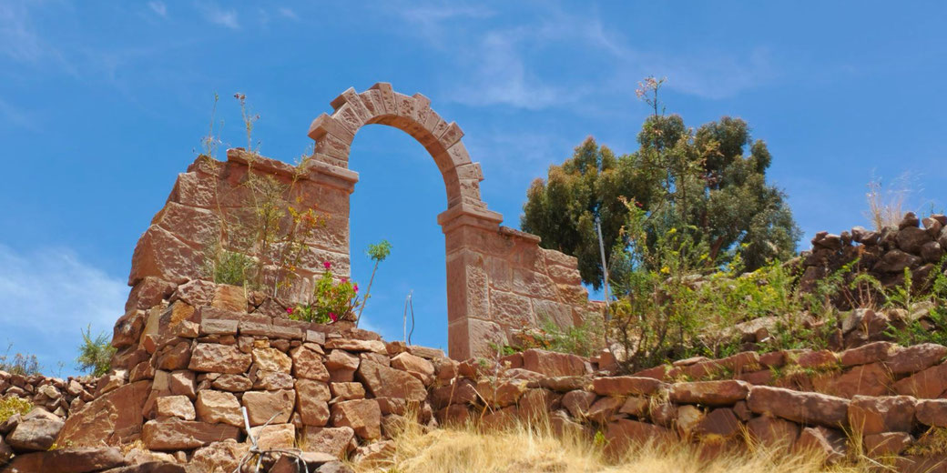 A rustic stone arch atop a rocky hillside, surrounded by dry grass and sparse vegetation, under a bright blue sky. It appears to be an ancient or traditional structure.