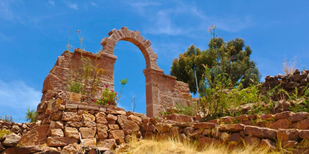 A rustic stone arch atop a rocky hillside, surrounded by dry grass and sparse vegetation, under a bright blue sky. It appears to be an ancient or traditional structure.