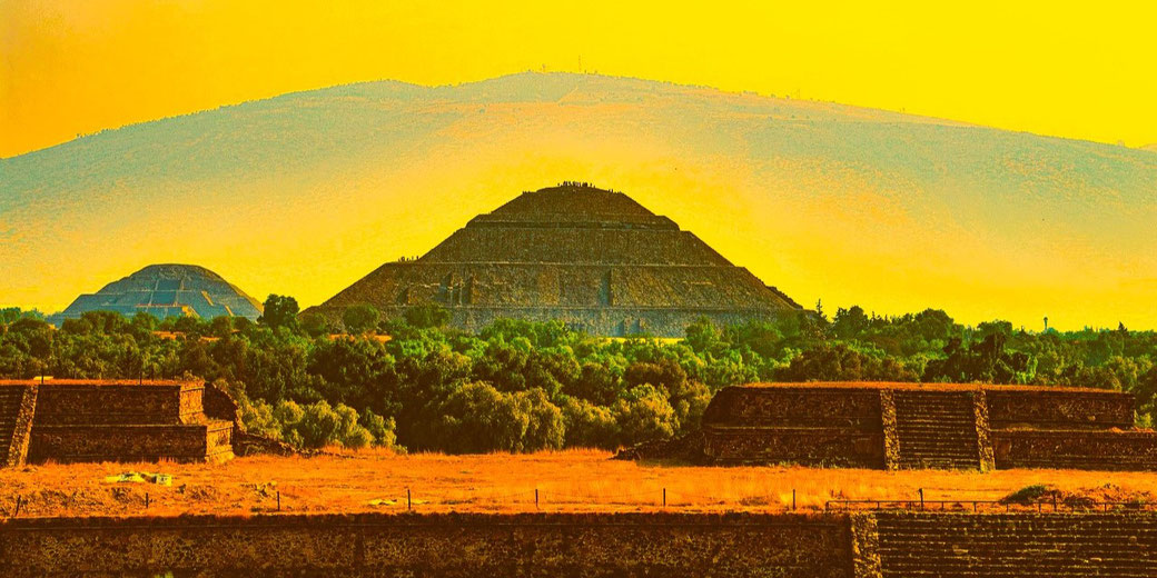 Pyramid of the Sun in Teotihuacan, Mexico, with the Pyramid of the Moon in the background. The golden-hued sky and mountainous backdrop enhance the scene's dramatic appeal.