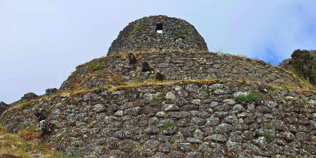 Ancient stone beehive structure on a grassy hillside beneath an overcast sky, viewed from below with rough masonry detail.