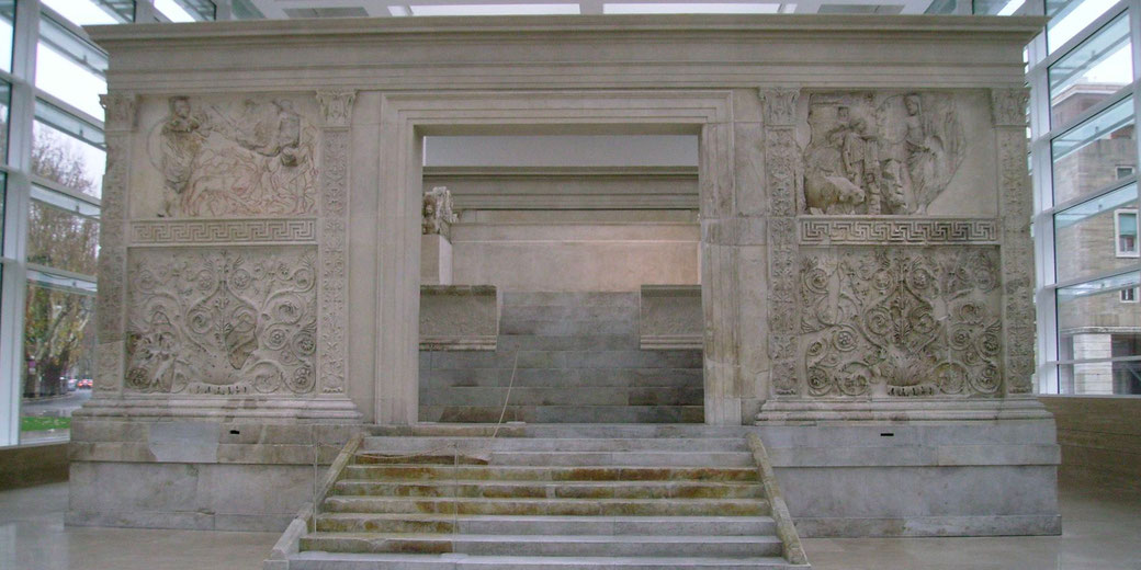 Large marble Roman altar with carved reliefs and floral motifs, centered by a stairway leading into an enclosed structure.