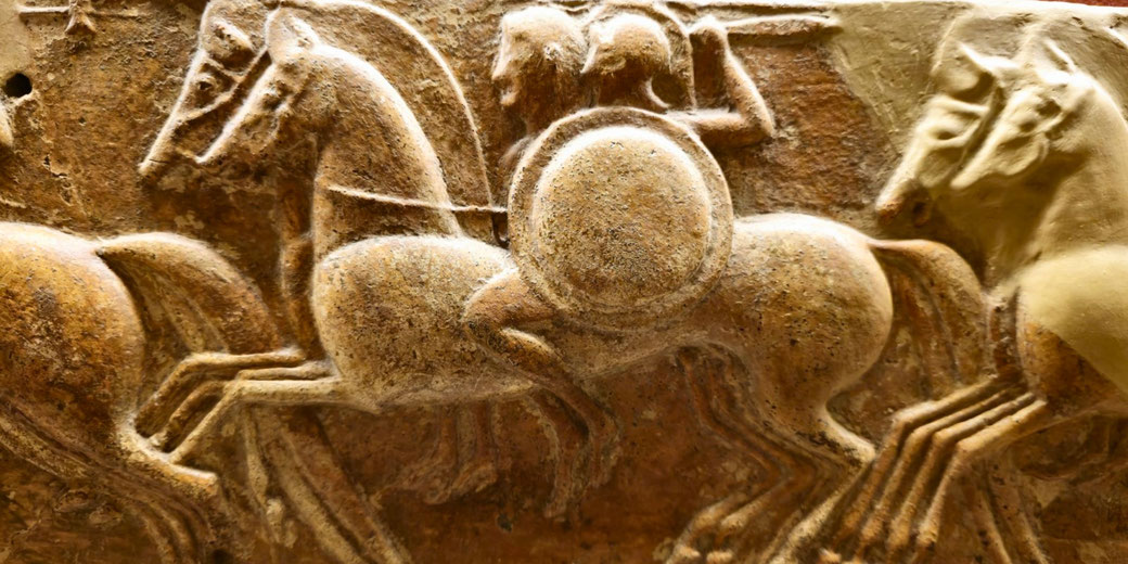Ancient relief carving shows warriors on horseback, one holding a spear and shield, in dynamic motion with raised hooves and detailed musculature.