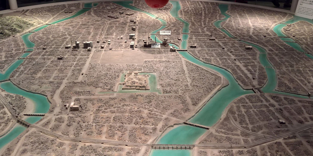 A detailed scale model shows Hiroshima after the atomic bombing, with rivers, ruins, bridges, and building remnants visible throughout the devastated city landscape.