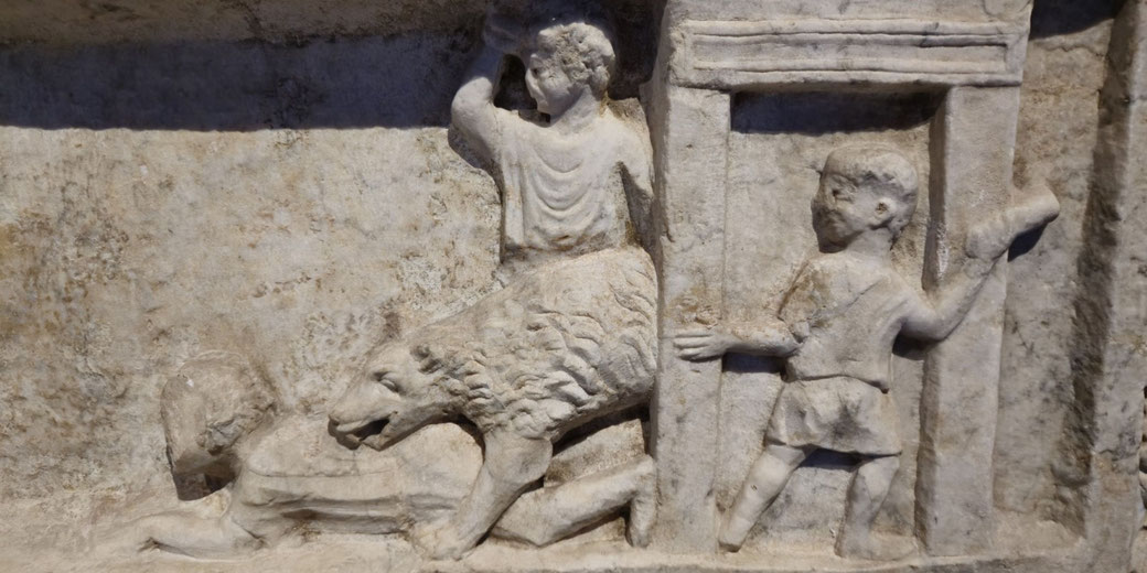 Ancient relief showing a wild animal attack: a beast lunges at a fallen figure, while two others look on, one holding a door-like structure, the other raising an arm in alarm.