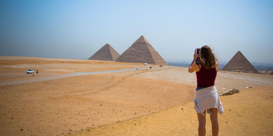 A tourist in a red top and white skirt takes a photo of the Pyramids of Giza under a clear sky. The vast desert landscape features a few cars and people, with Cairo's skyline in the distance.