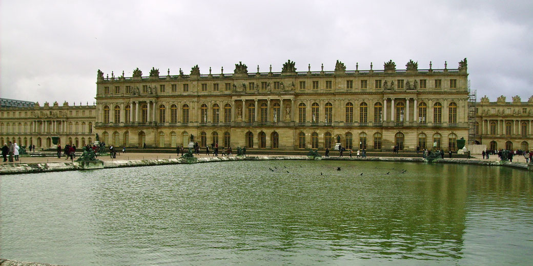 The Palace of Versailles stands behind a large reflecting pool with tourists walking along its edge.